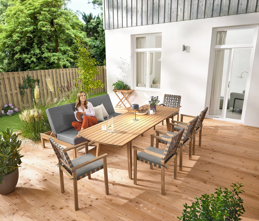 Žena sedí na zahradní lavici 2 v 1 ‒ dvojité lehátko. Na rozkládacím zahradním stole se zaoblenými čely, cca 180‒240 cm, stojí šedá solární stolní lampa. K dispozici je také zahradní odkládací stolek a stohovatelné zahradní židle s pásovým výpletem.