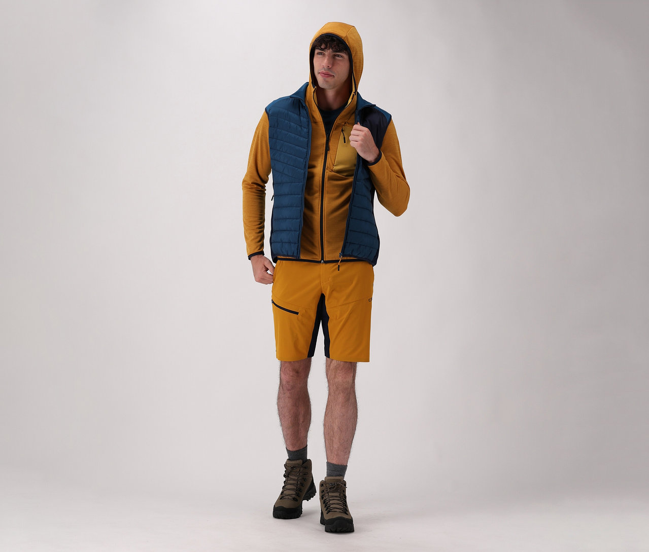 Muž na světlém pozadí má na sobě pánskou bundu s kapucí CMP, pánskou hybridní vestu CMP, šortky a boty.