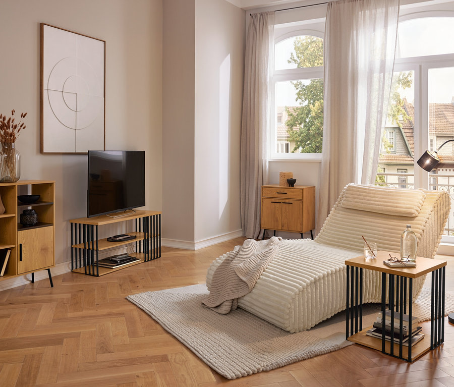 Moderní obývací pokoj s krémovou lenoškou na světlém koberci, dřevěnou skříňkou, TV stolkem a odkládacím stolkem. Velká okna se závěsy s výhledem ven.