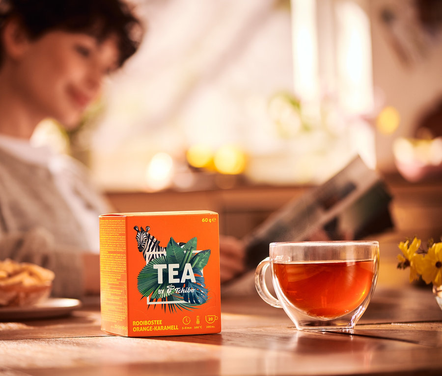 Žena čte noviny u stolu s šálkem čaje a balením čaje Rooibos TEA by Tchibo s příchutí pomeranč/karamel.