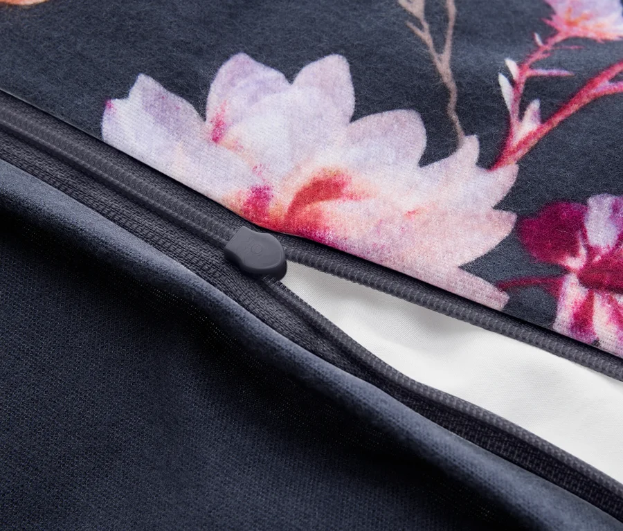 Detail tmavě šedé látky s květinovým vzorem s růžovými a fialovými květy, částečně rozepnuté, odhalující bílou podšívku. Je vidět šedý jezdec zipu.