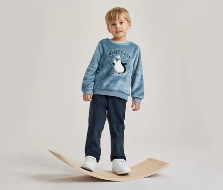 Malý chlapec stojí na dřevěné balanční desce. Má na sobě modrý top s dlouhým rukávem s grafikou tučňáka a nápisem „WINTER FUN“, tmavě modré manšestrové kalhoty a bílé tenisky.