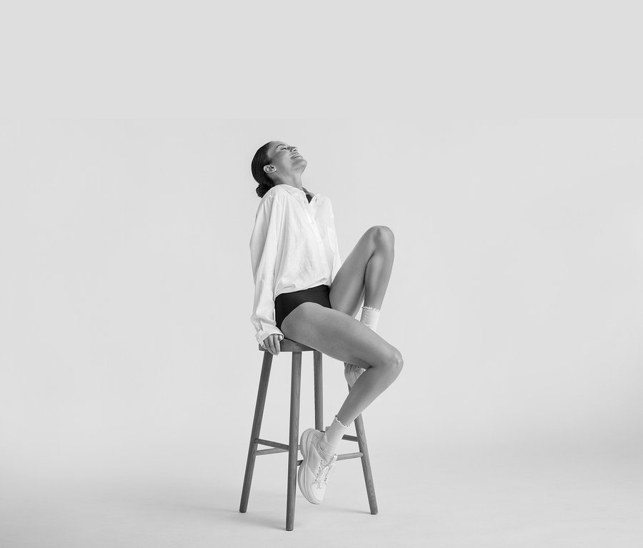 Žena sedí usměvavá na stoličce, má na sobě bílou Dlouhá lněná halenka, černé spodní prádlo, bílé ponožky a bílé tenisky.