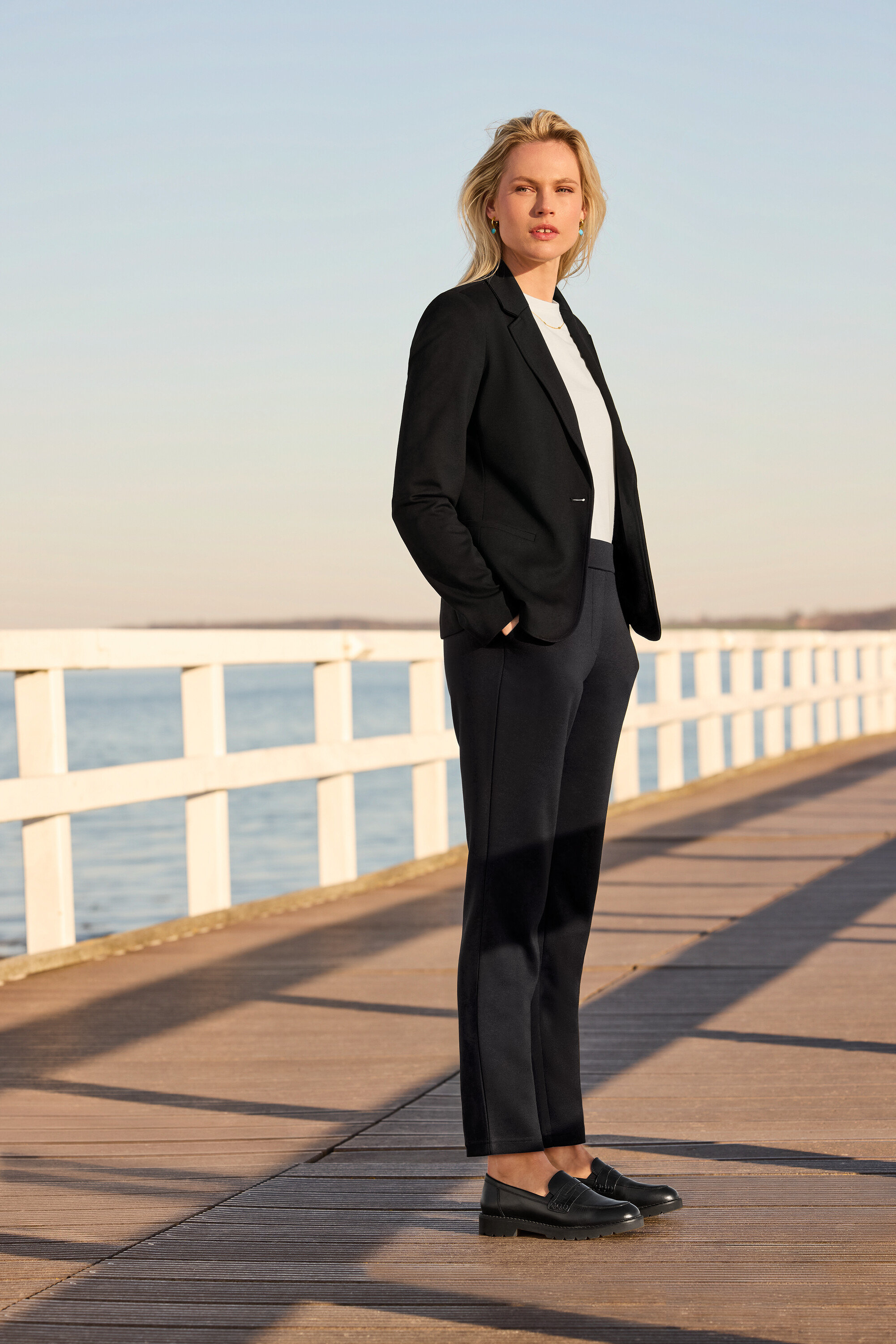 Žena stojící na molu má na sobě triko Basic, bílé, kalhoty joggpant, žerzejový blejzr a kožené mokasíny. Má náhrdelník a náušnice s přívěskem s kamínkem.