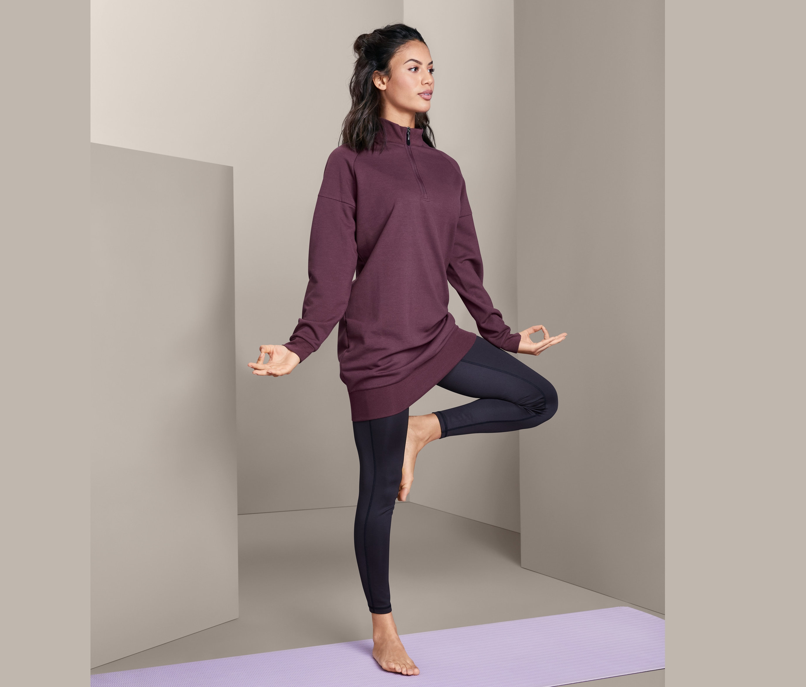 Žena v dlouhém triku na jógu stojí na fialové podložce na jógu a dělá pozici stromu.