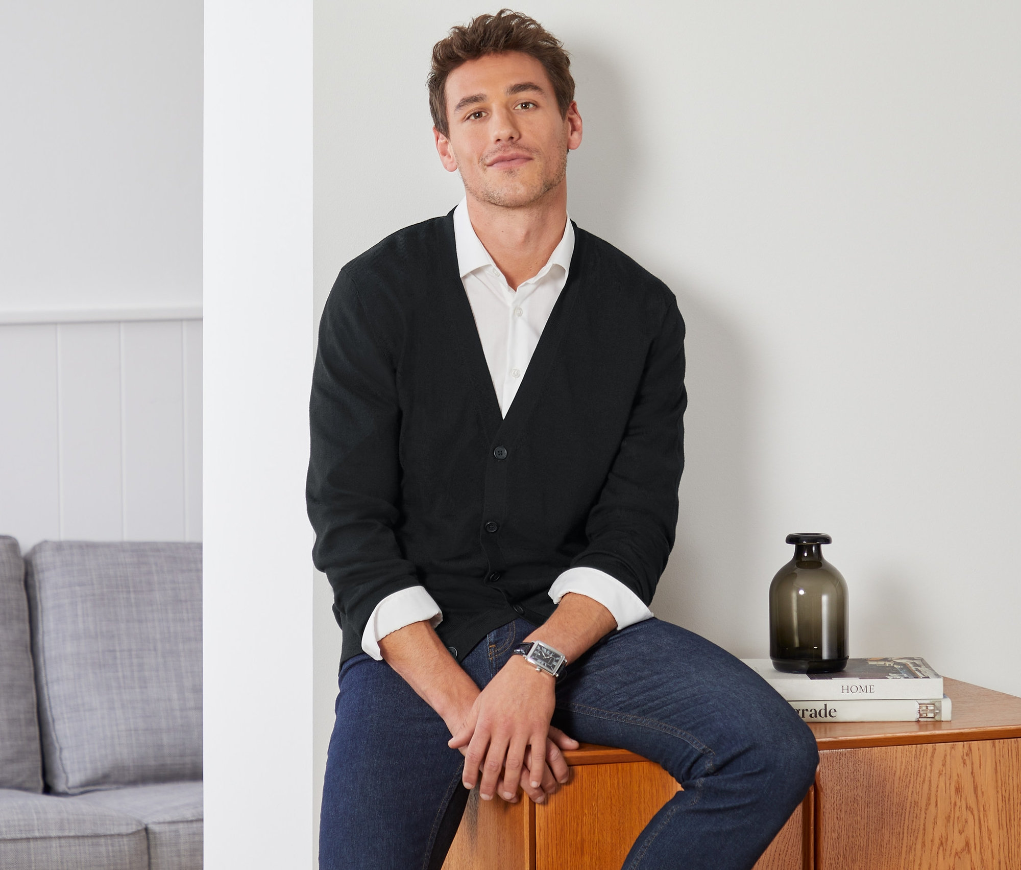 Muž sedí na dřevěné skříňce a má na sobě černý vlněný kardigan.