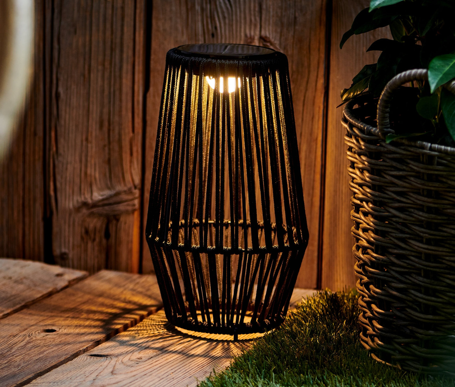 Černý solární lampáš stojí na dřevěné palubě vedle proutěného koše s rostlinou.