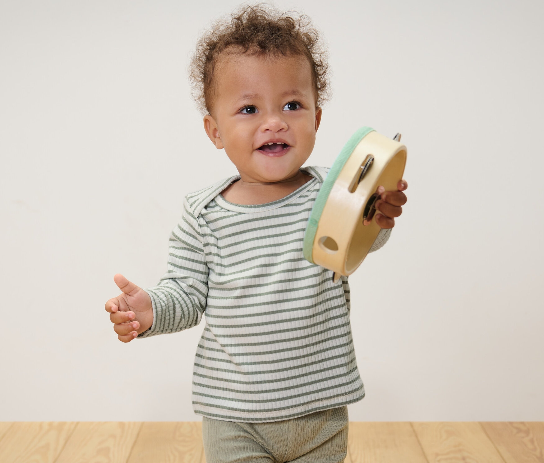 Dítě stojí a drží tamburínu. Má na sobě světle modré a zelené tričko s dlouhým rukávem a žebrovanou strukturou.