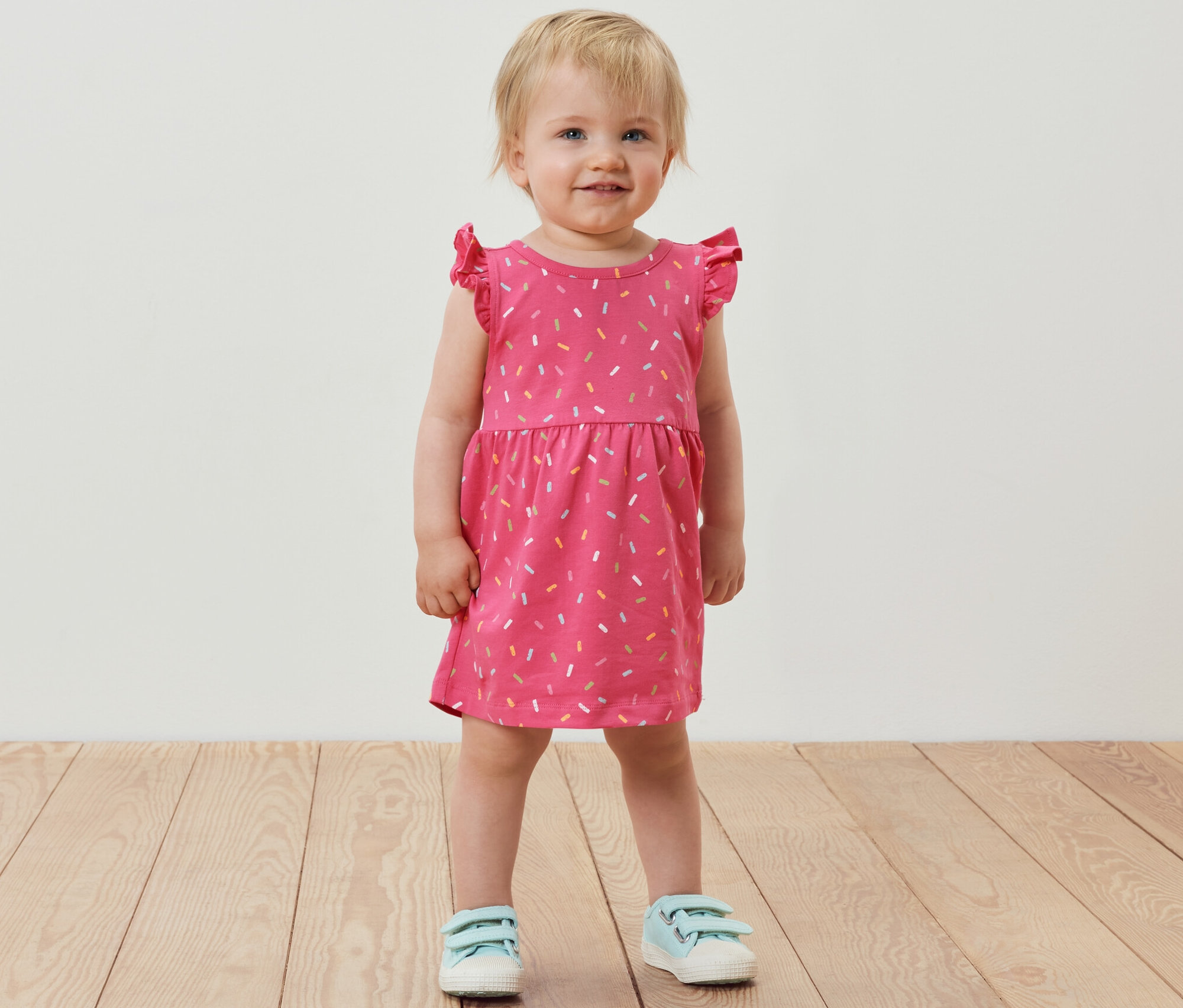 Usmívající se holčička stojí a má na sobě růžové žerzejové šaty s krátkými rukávy.