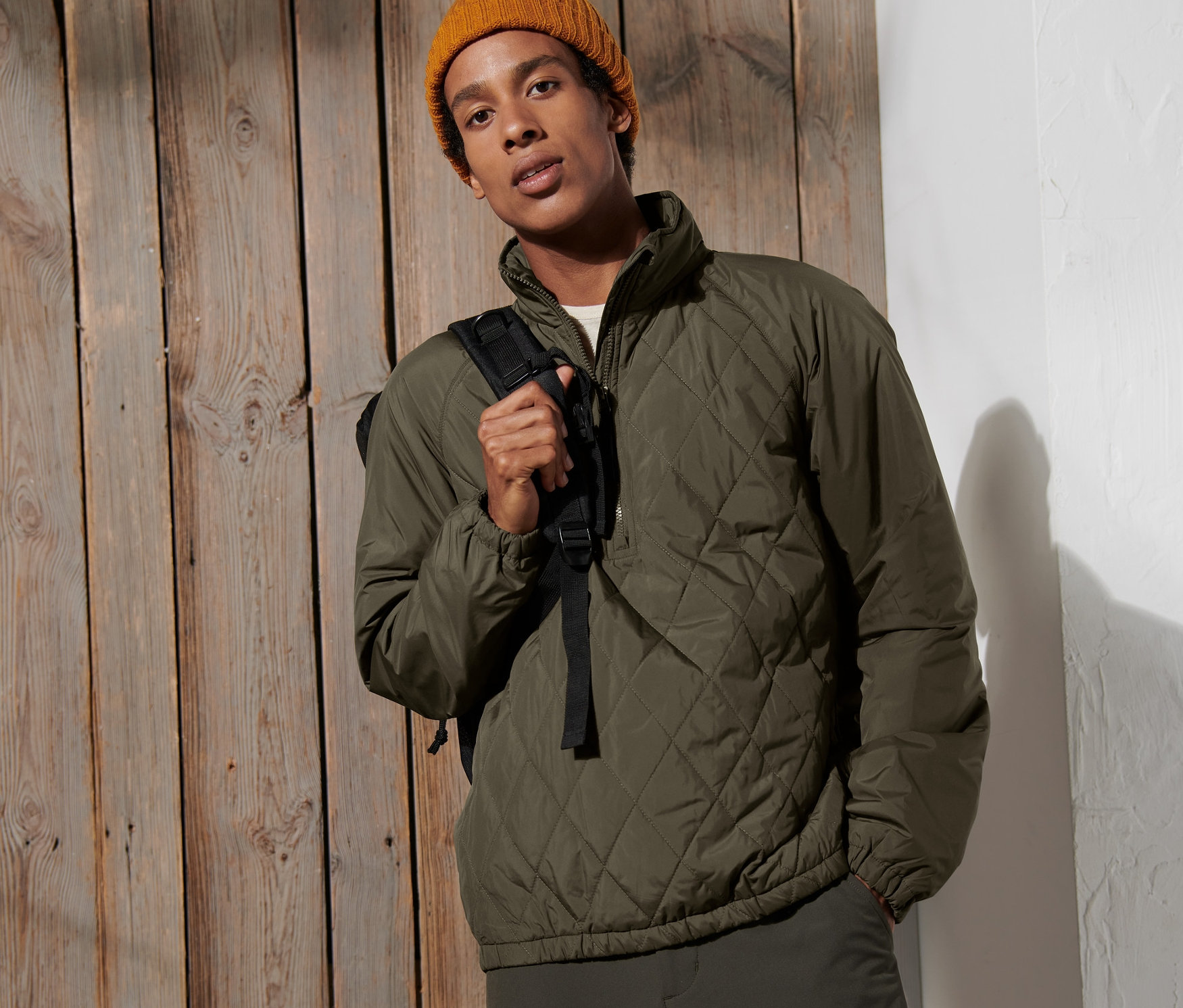 Muž pózuje na pozadí dřevěné stěny a má na sobě větrovku a žlutou čepici.