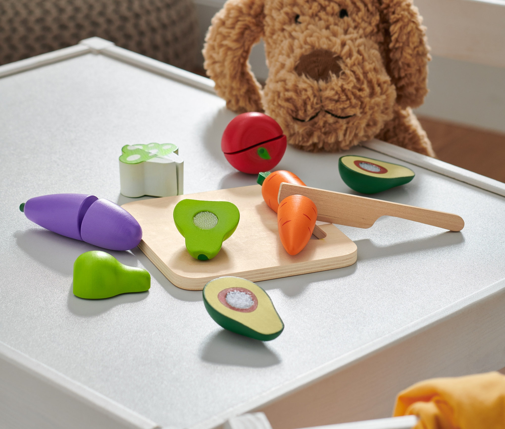 Plyšový pes sedí na bílém stole vedle dřevěných hraček ovoce a zeleniny.