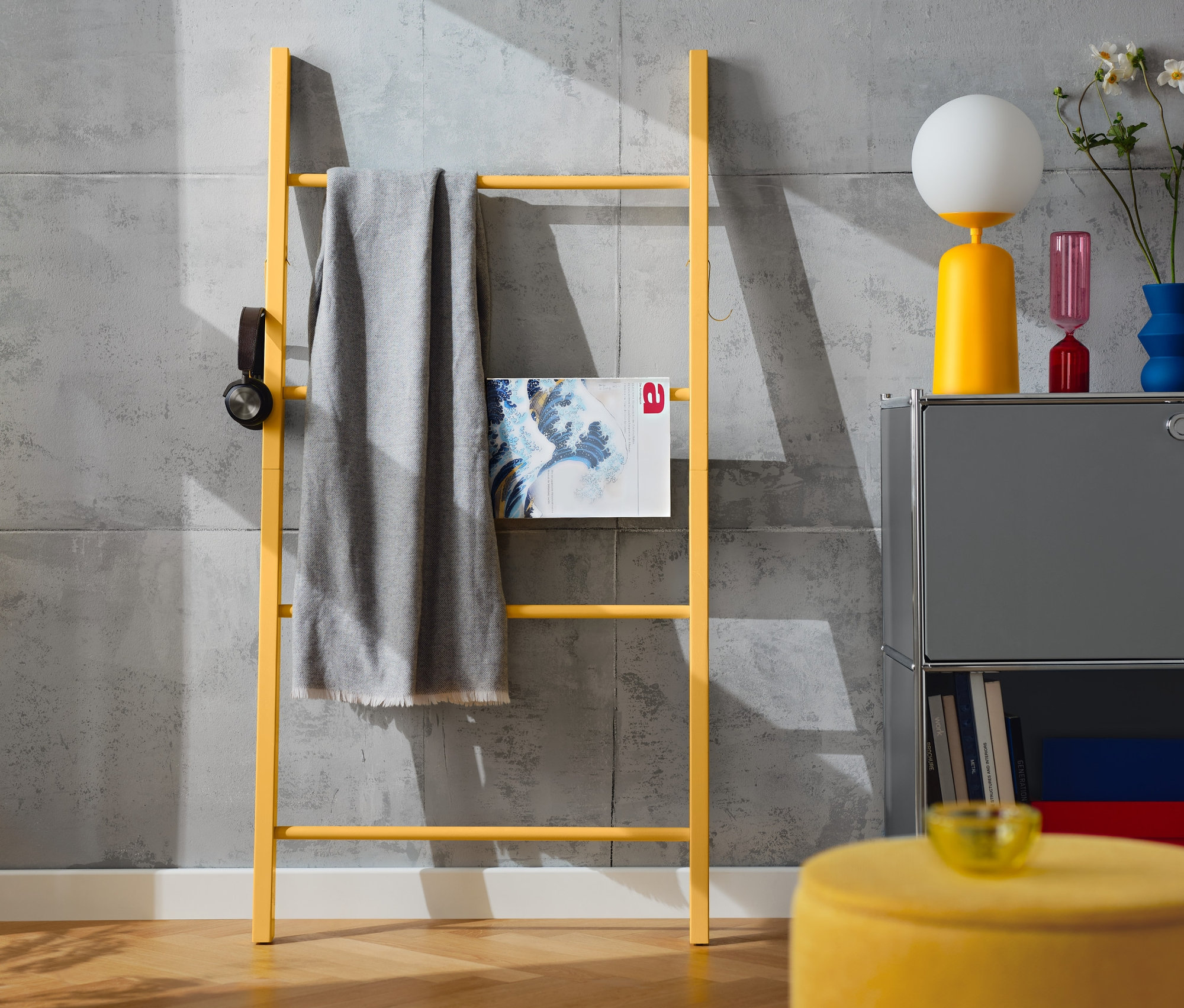 Žlutý dřevěný žebřík s háčky, na kterém visí šedá deka. Vedle žebříku stojí žlutá stolní lampa se skleněnou koulí.