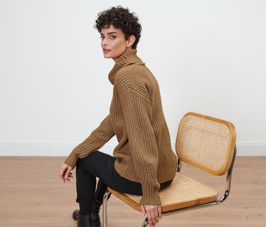 Žena sedí na židli a má na sobě hnědý svetr z hrubé pleteniny s rolákovým límcem.