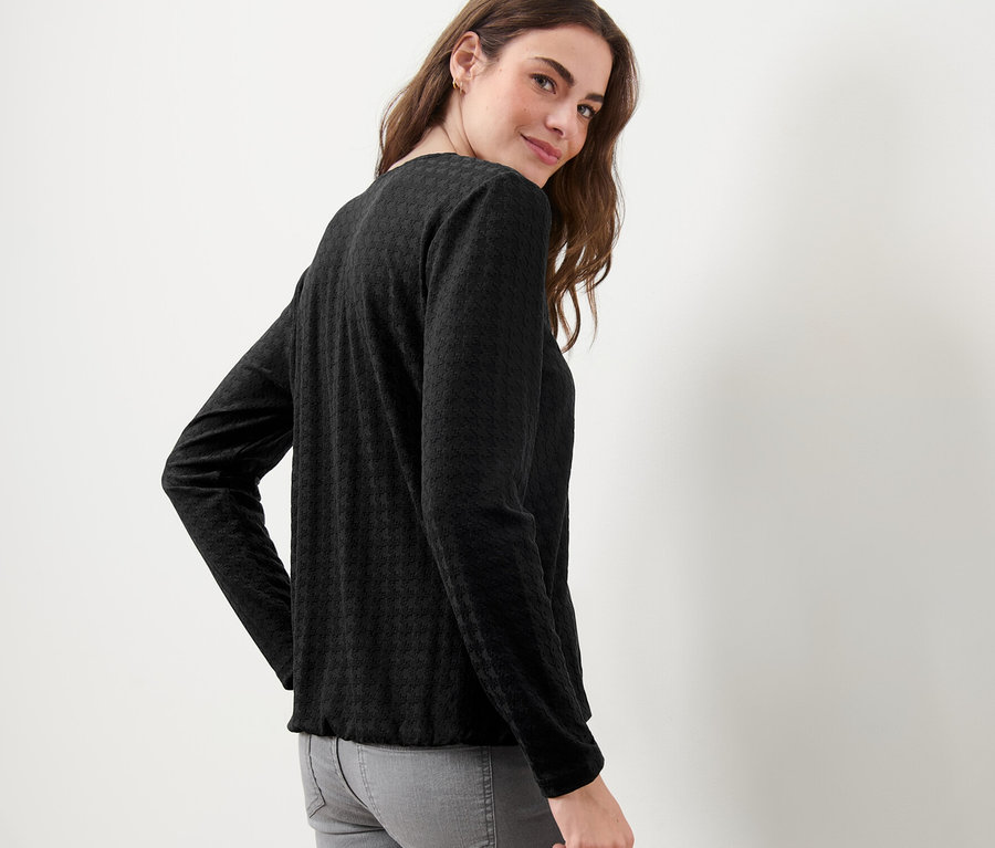 Žena se dívá přes rameno a má na sobě černé strukturované triko s dlouhým rukávem.