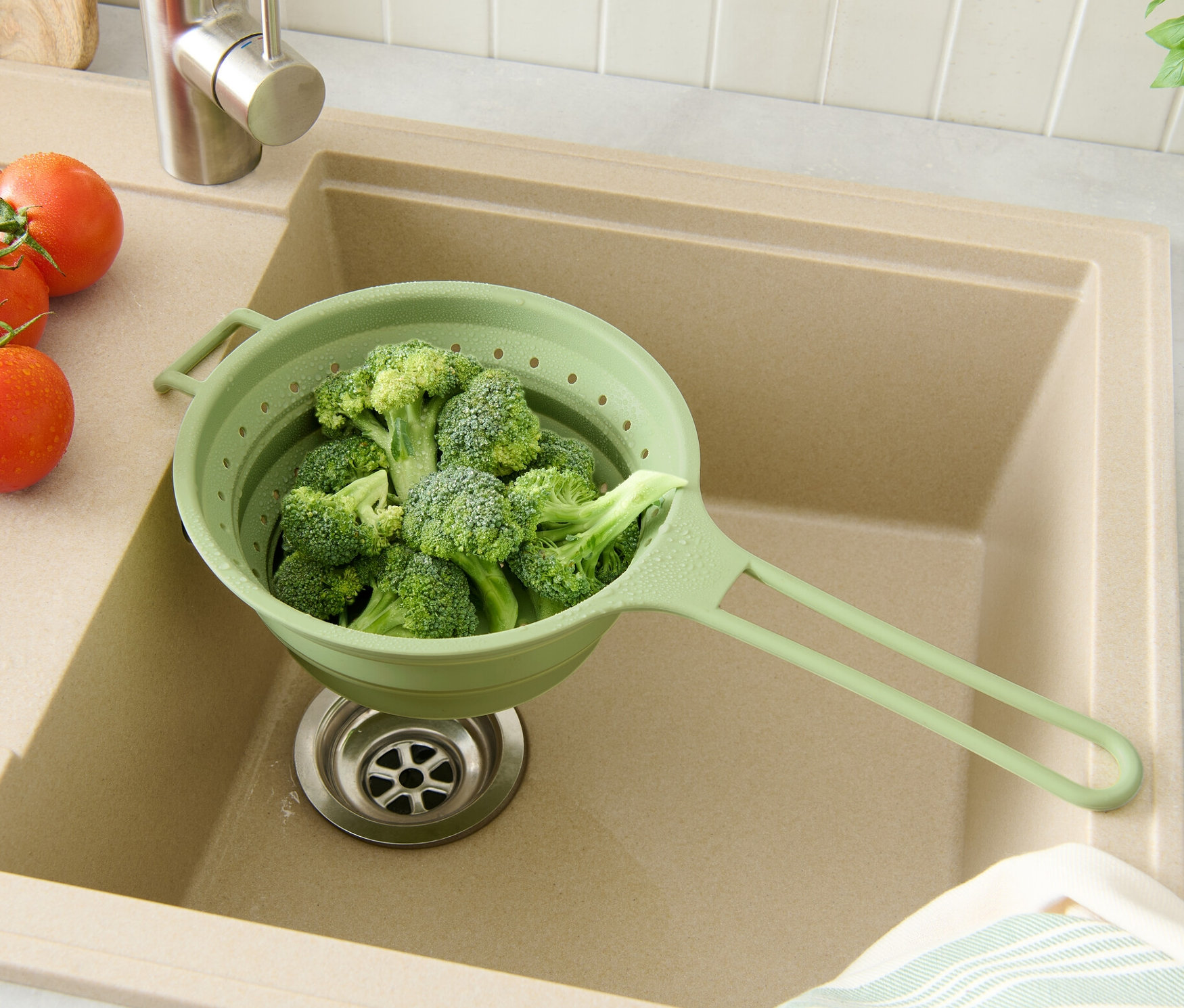 Brokolice v zeleném prostorově úsporném cedníku v béžovém dřezu s rajčaty a bylinkami.