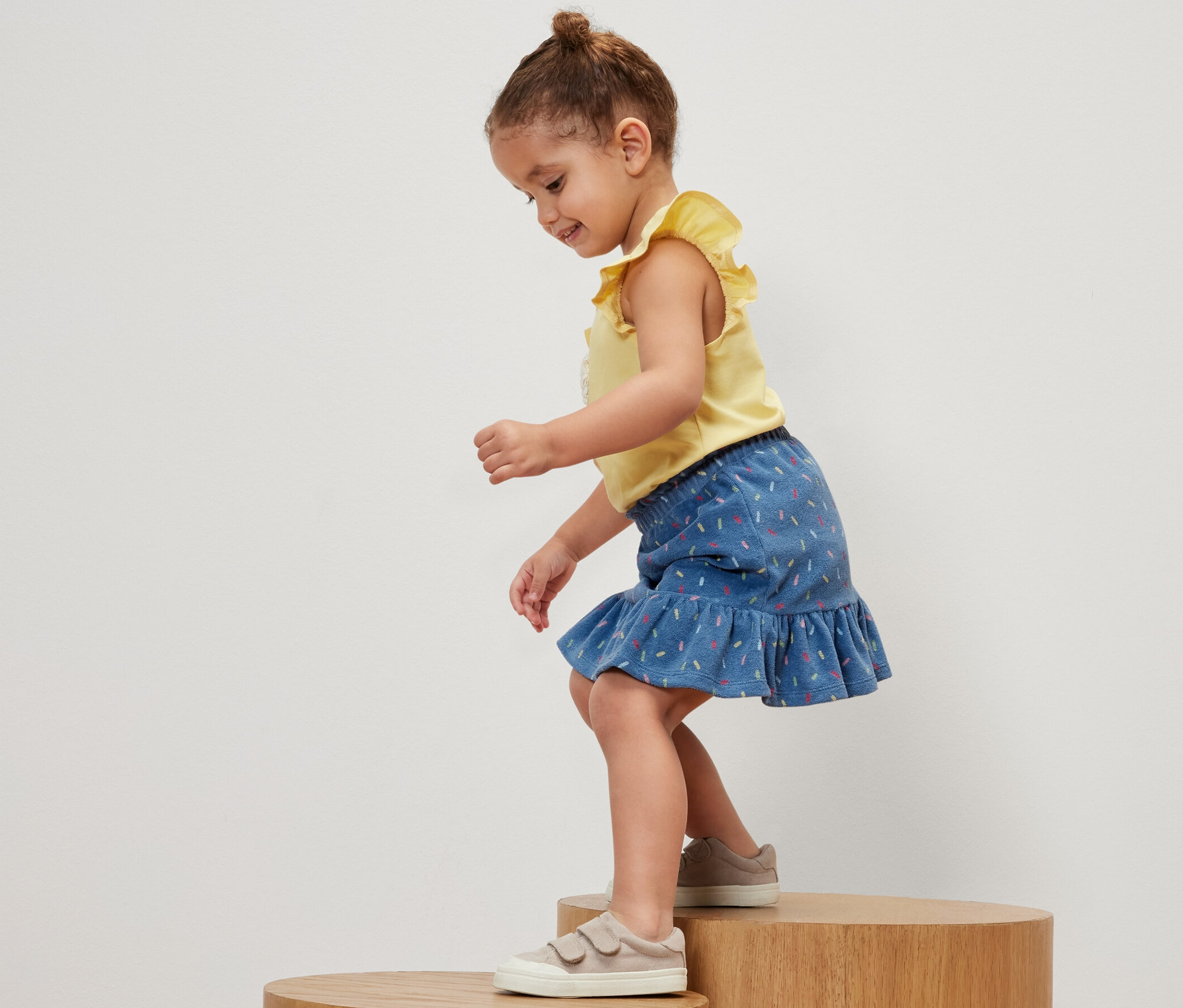 Holčička stojí na dřevěném podstavci a má na sobě žluté tričko a dětskou froté sukni.