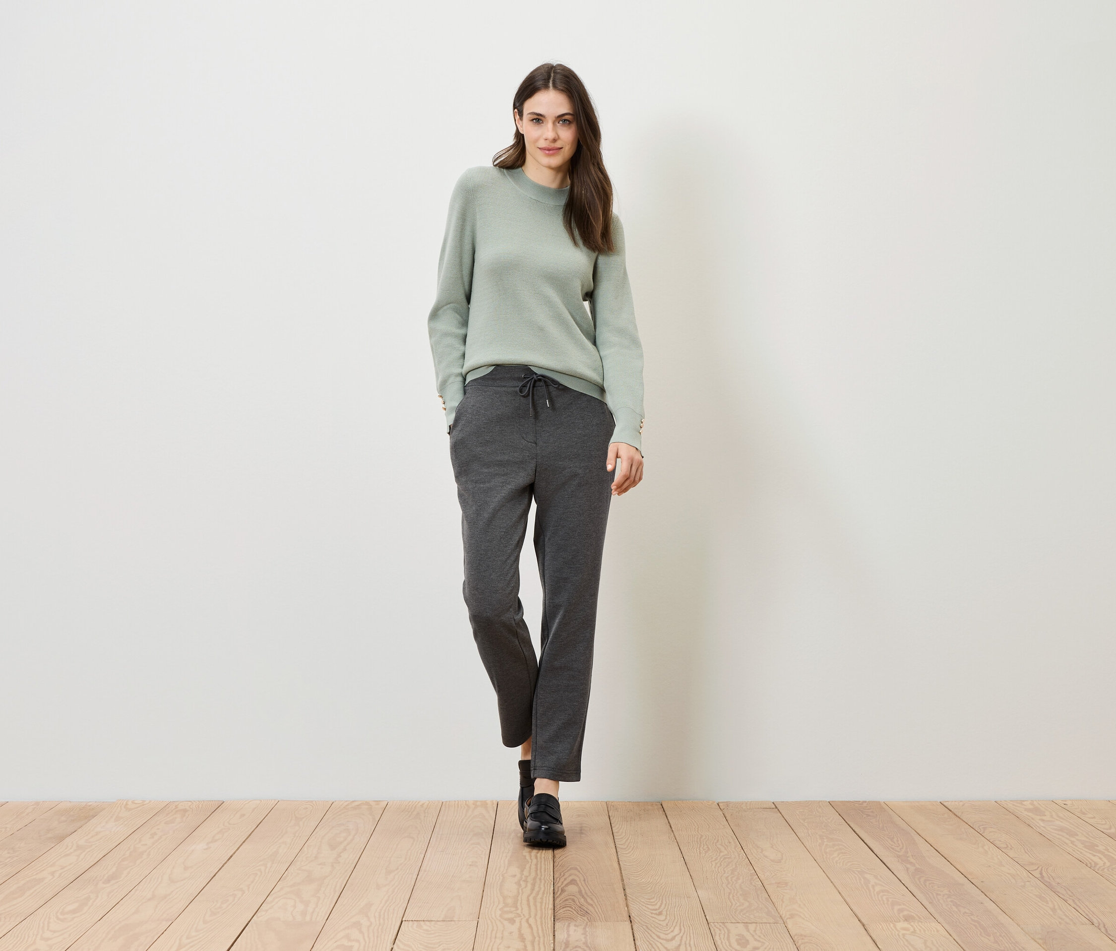 Žena stojí na dřevěné podlaze na pozadí bílé stěny. Má na sobě zelený svetr a šedé kalhoty.