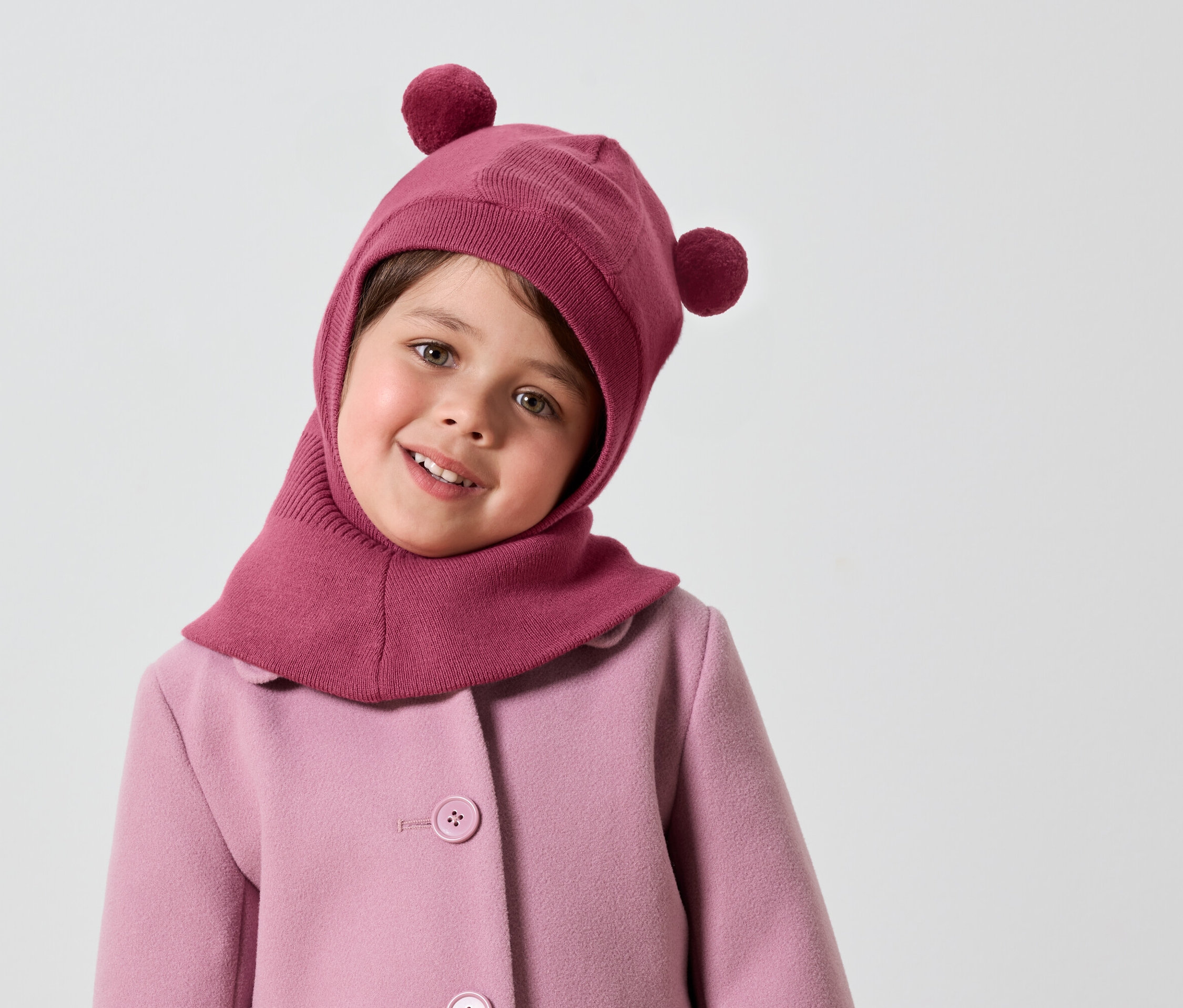 Usmívající se holčička v růžové kukle s bambulkami a růžovém kabátě.