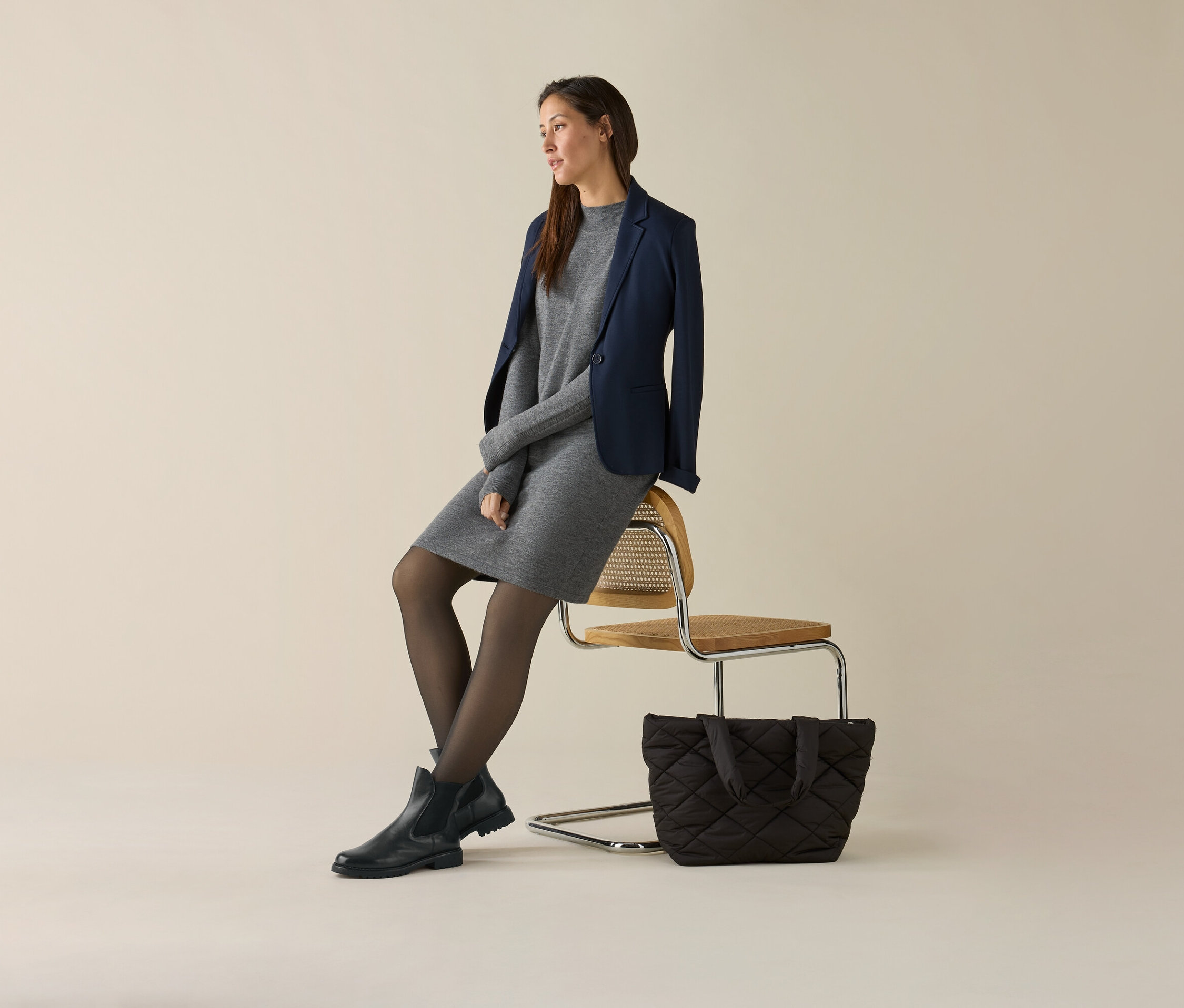 Žena sedí na židli a má na sobě šedé šaty a tmavě modré žerzejové sako. Vedle židle je kabelka.