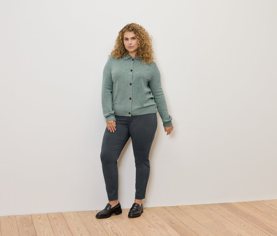 Žena s kudrnatými vlasy stojí a má na sobě zelený svetr, šedé kalhoty a černé mokasíny.