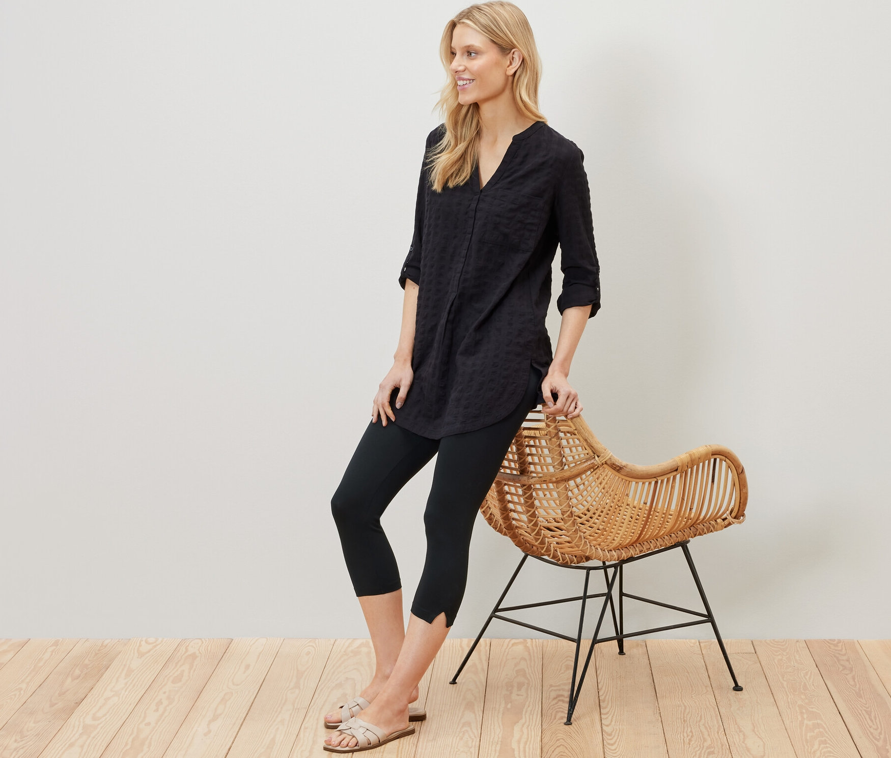 Blondýna se opírá o proutěné křeslo a má na sobě černé 3/4 legíny s detaily.