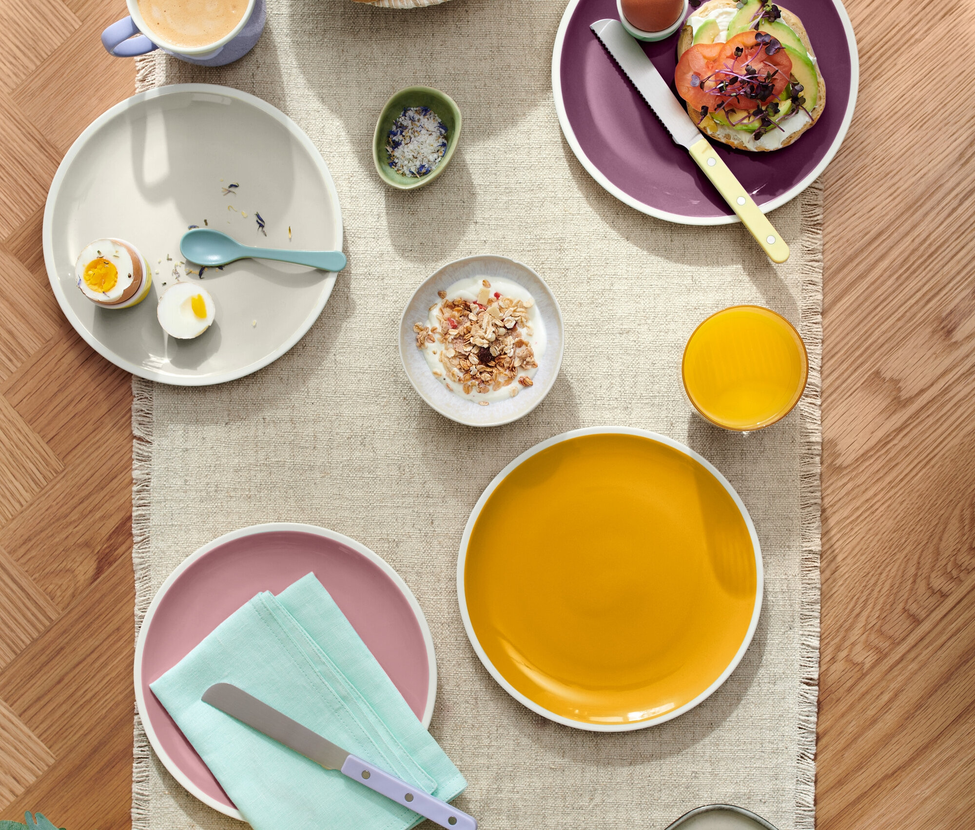 Pohled shora na prostřený stůl s různými pokrmy, včetně pečiva v košíku, talířů s vejci, avokádovým toastem a sýrem, spolu s nápoji, jako je káva a džus.