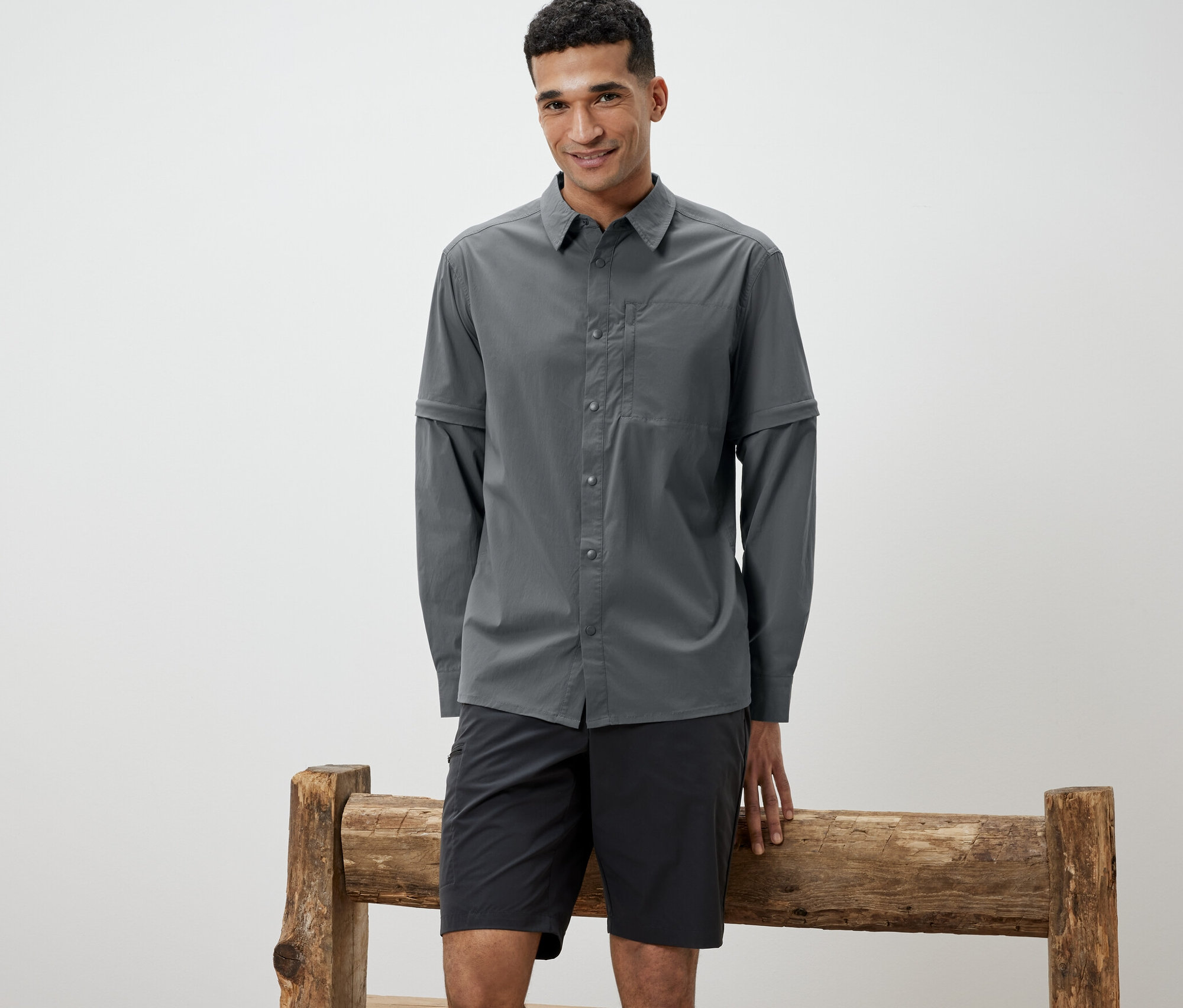 Muž opřený o dřevěnou lavici má na sobě šedou outdoorovou košili a černé šortky.