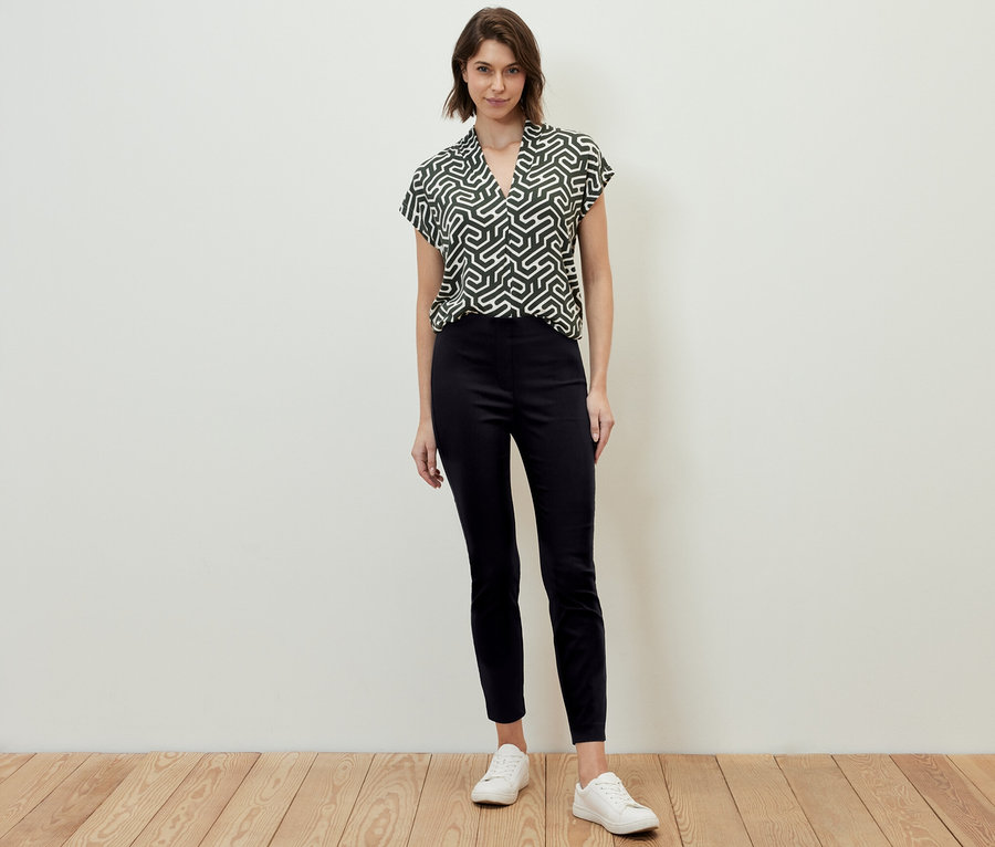 Žena stojí a má na sobě černé strečové kalhoty a vzorovanou blůzu.