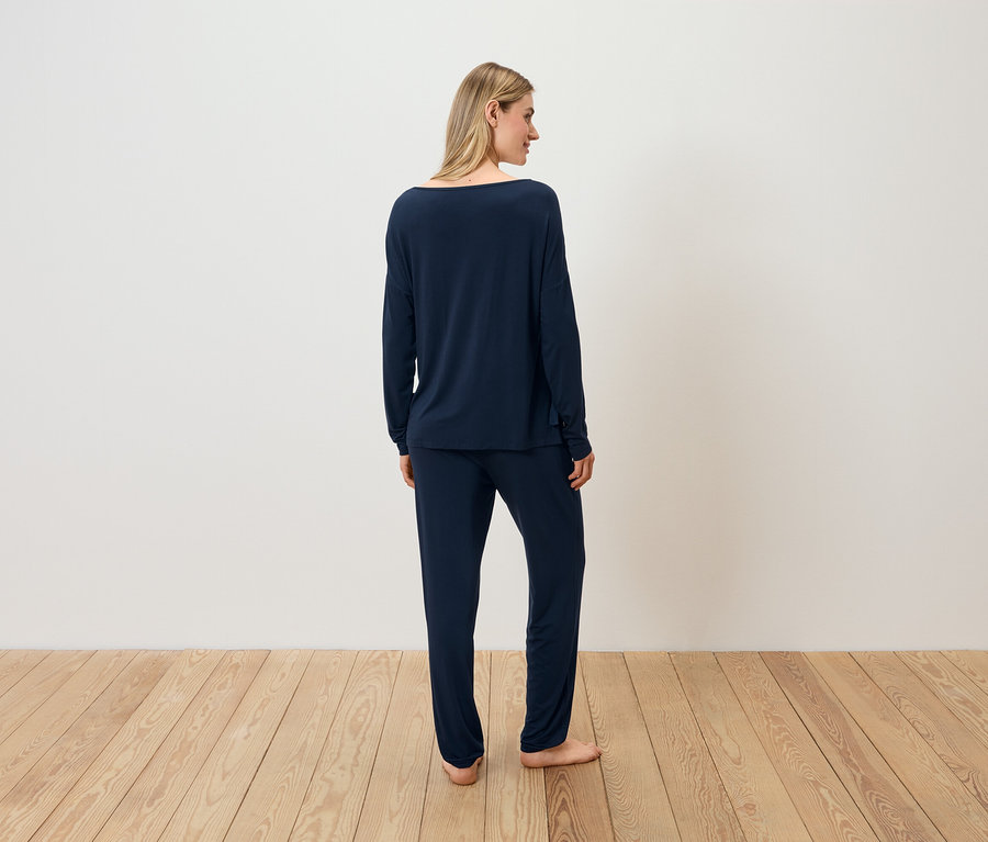 Žena v tmavě modrém pyžamu stojí zády ke kameře na dřevěné podlaze.