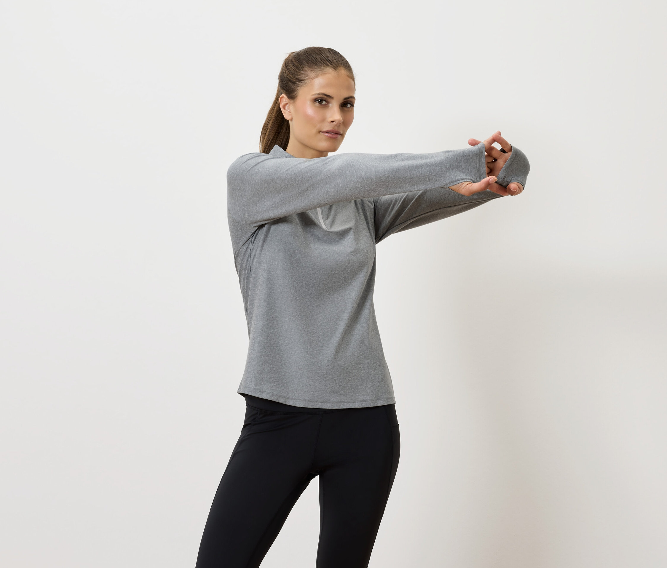 Žena protahuje ruce v šedé košili s dlouhým rukávem a černých legínách.