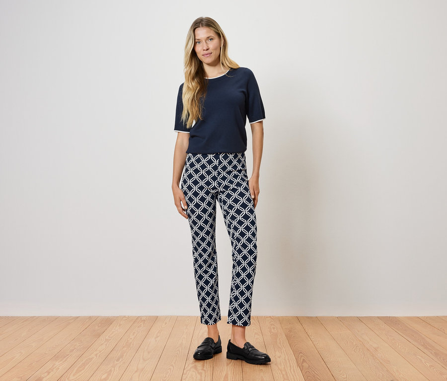 Žena stojí na dřevěné podlaze a má na sobě tmavě modré tričko a vzorované kalhoty.