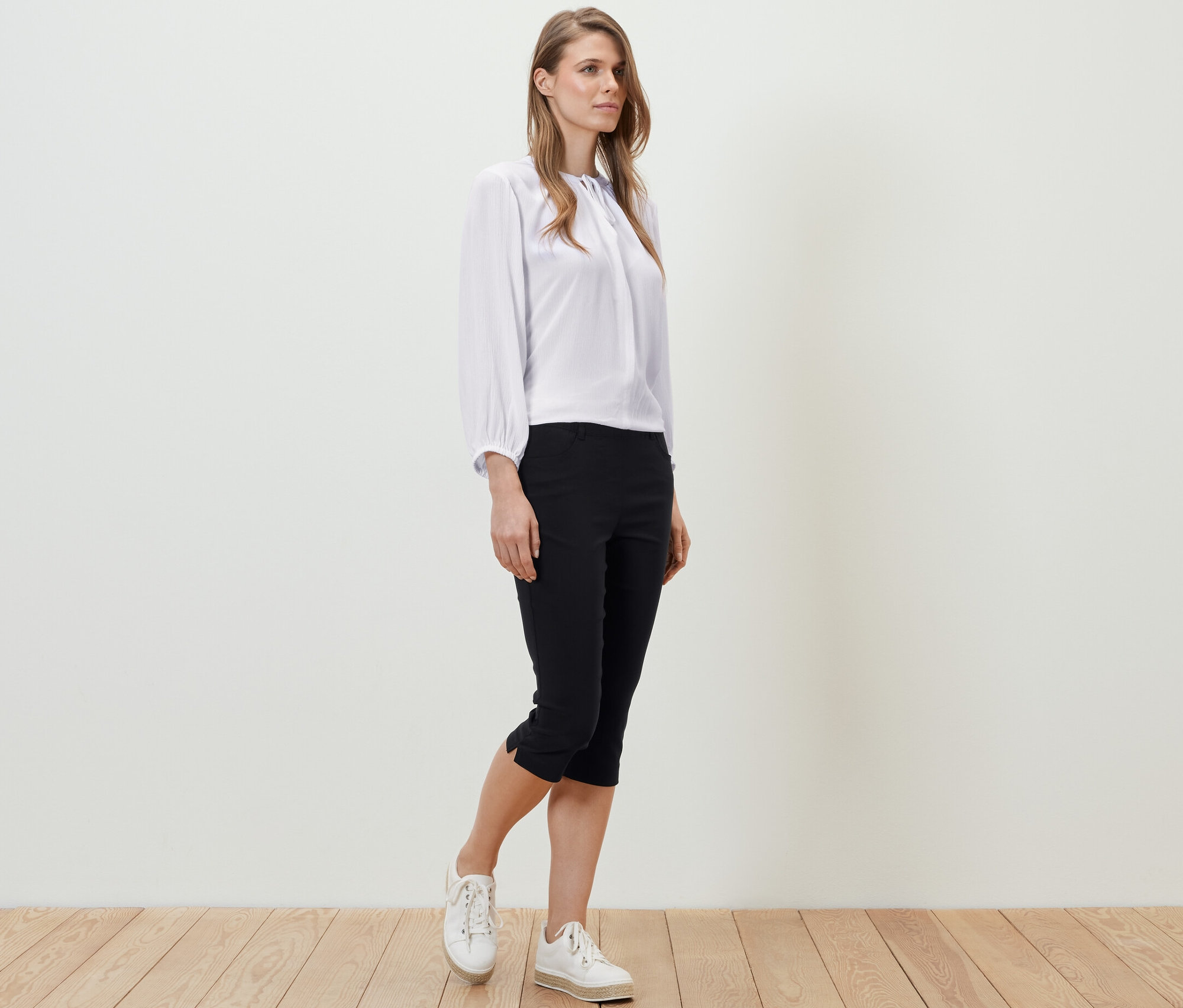 Žena stojící na dřevěné podlaze má na sobě bílou blůzu a černé 3/4 bengalínové kalhoty.