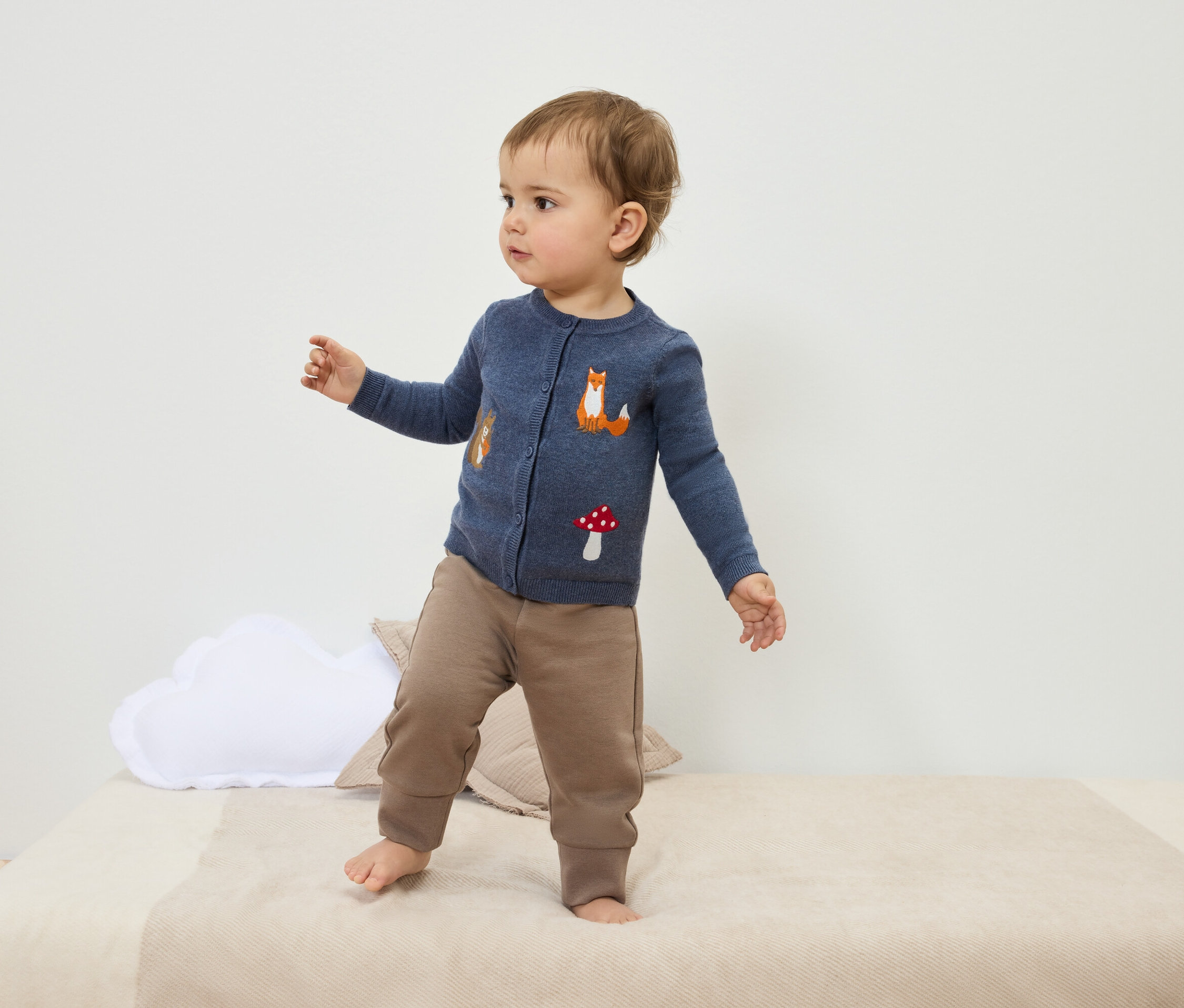 Batole stojí na posteli v modrém svetru s liškou a houbou a hnědých kalhotách.