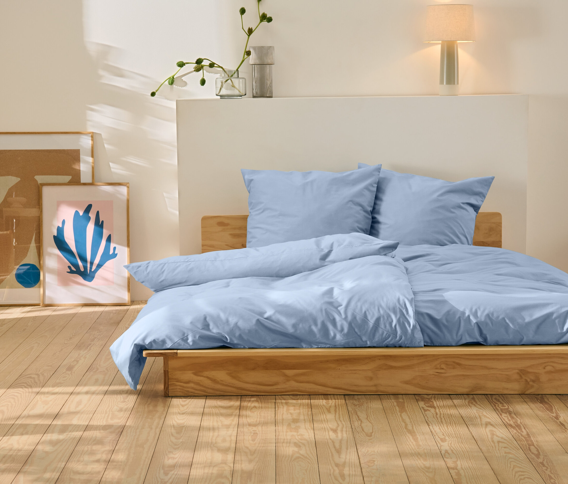 Postel s modrým povlečením stojí na dřevěné podlaze. Za postelí jsou dva obrazy.
