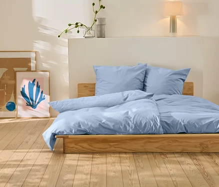 Postel s modrým povlečením stojí na dřevěné podlaze. Za postelí jsou dva obrazy.