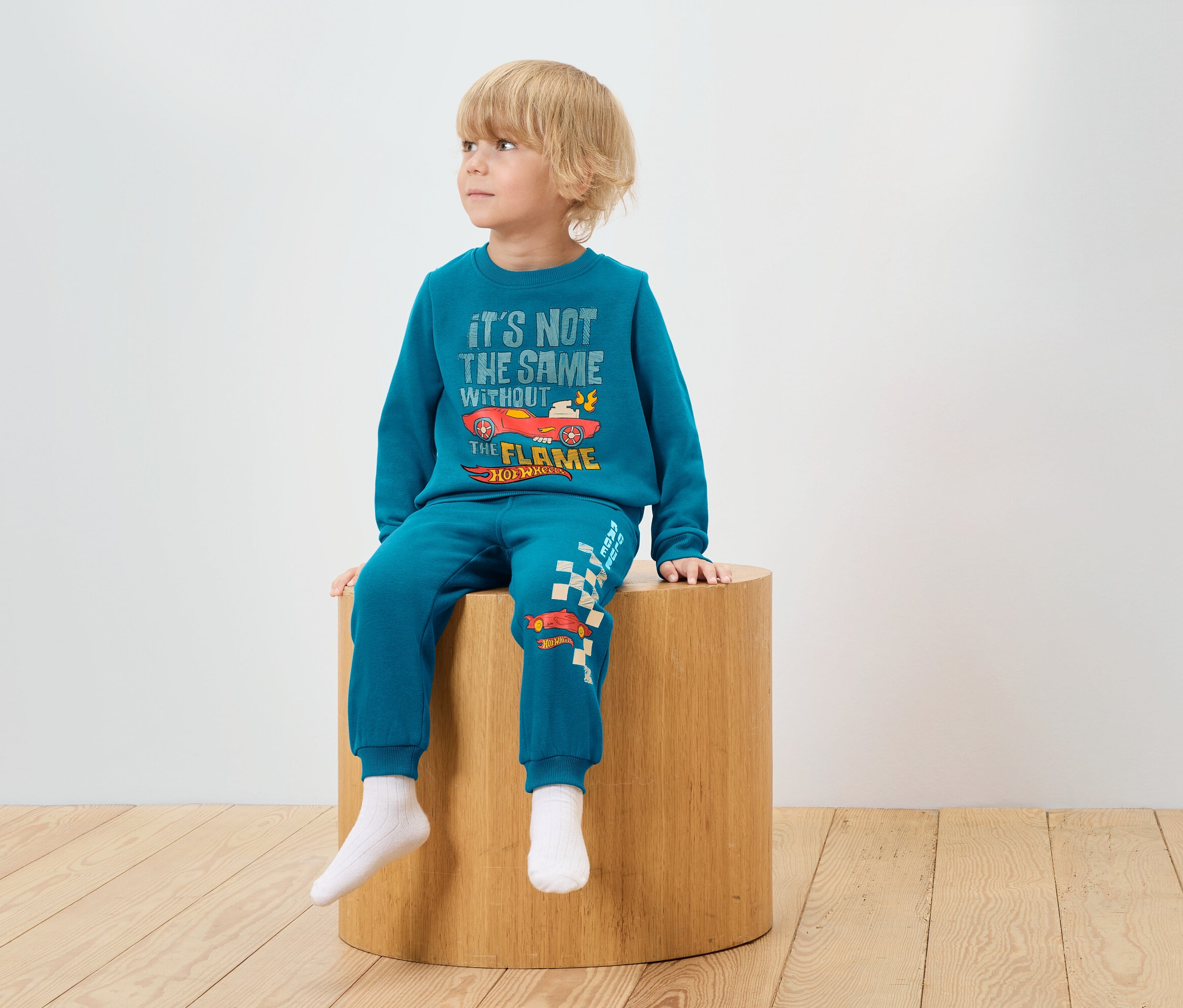 Chlapec sedí na dřevěné stoličce a má na sobě modrou soupravu s potiskem Hot Wheels.