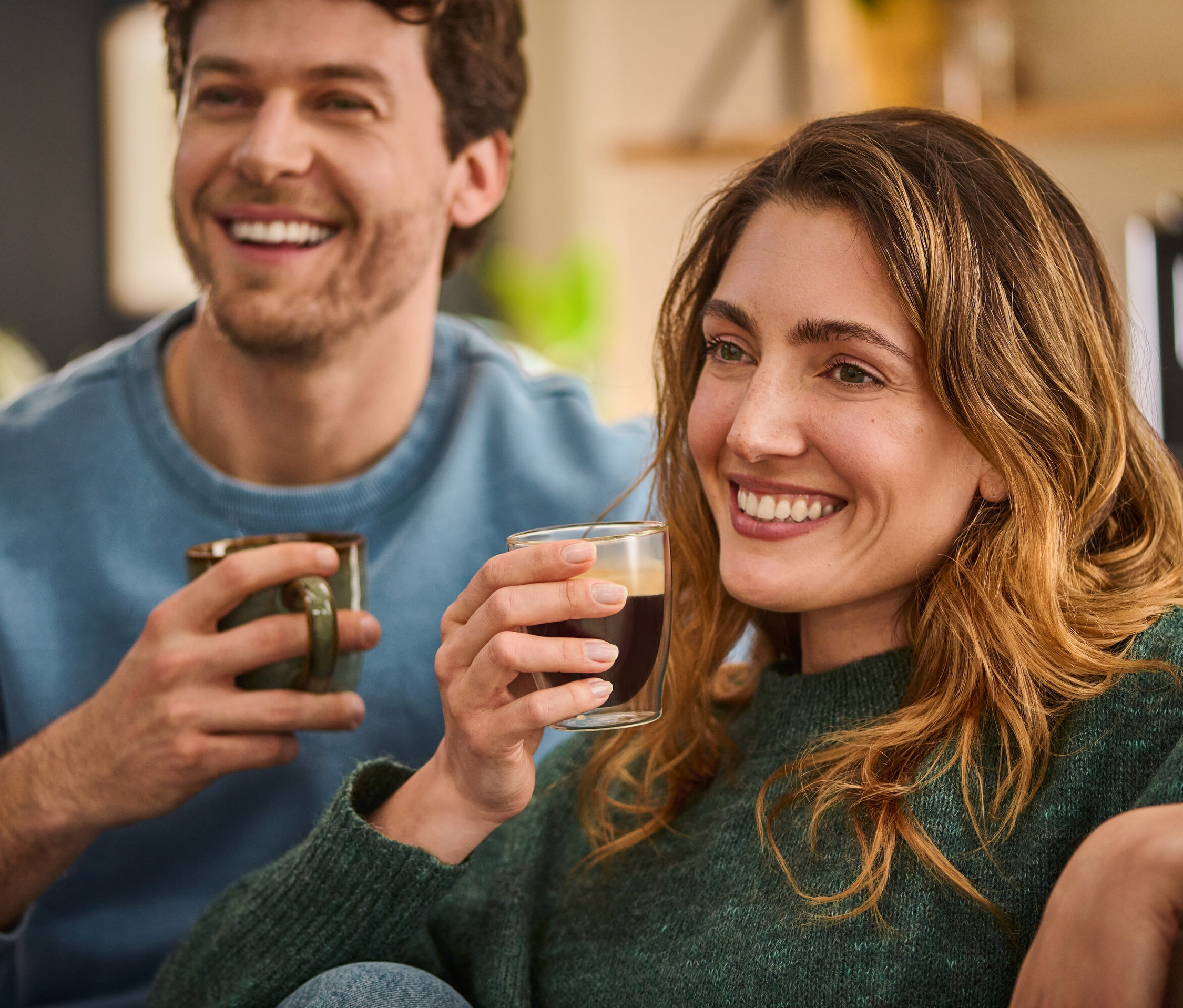 Usmívající se muž a žena sedí vedle sebe a drží šálky s kávou.