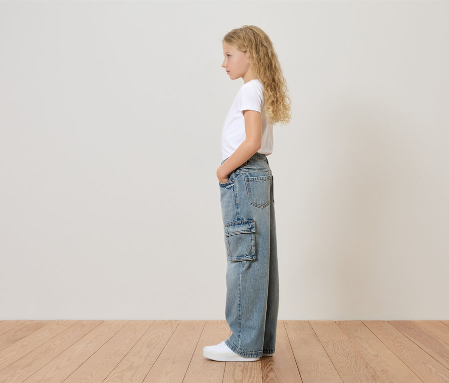Mladá dívka s blond kudrnatými vlasy stojí bokem a má na sobě bílé tričko, široké džíny s boční kapsou a bílé tenisky.