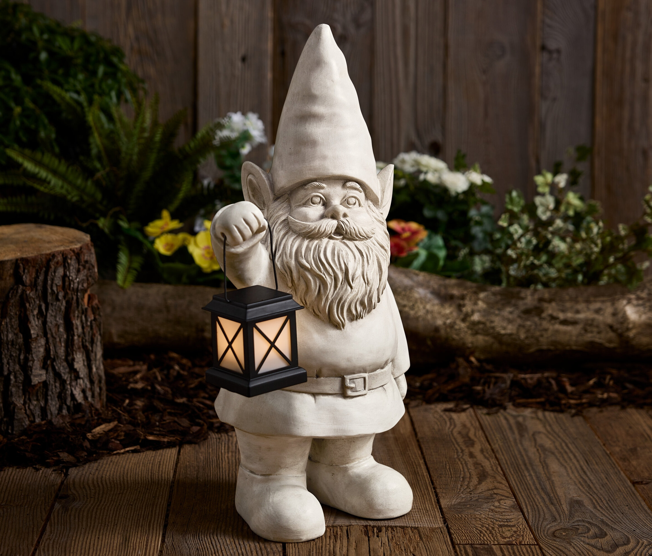 Kamenný zahradní trpaslík drží černou lucernu před dřevěným pozadím.