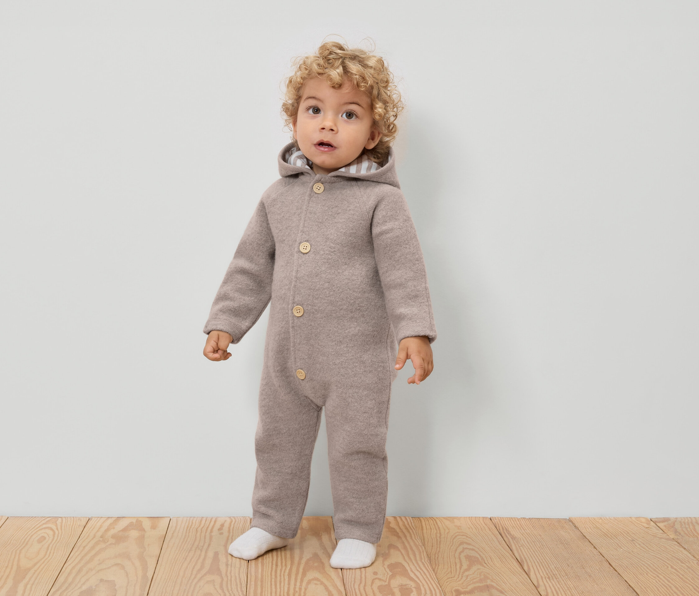 Malé dítě s kudrnatými vlasy má na sobě světle hnědou vlněnou kombinézu s kapucí a knoflíky a bílé ponožky.