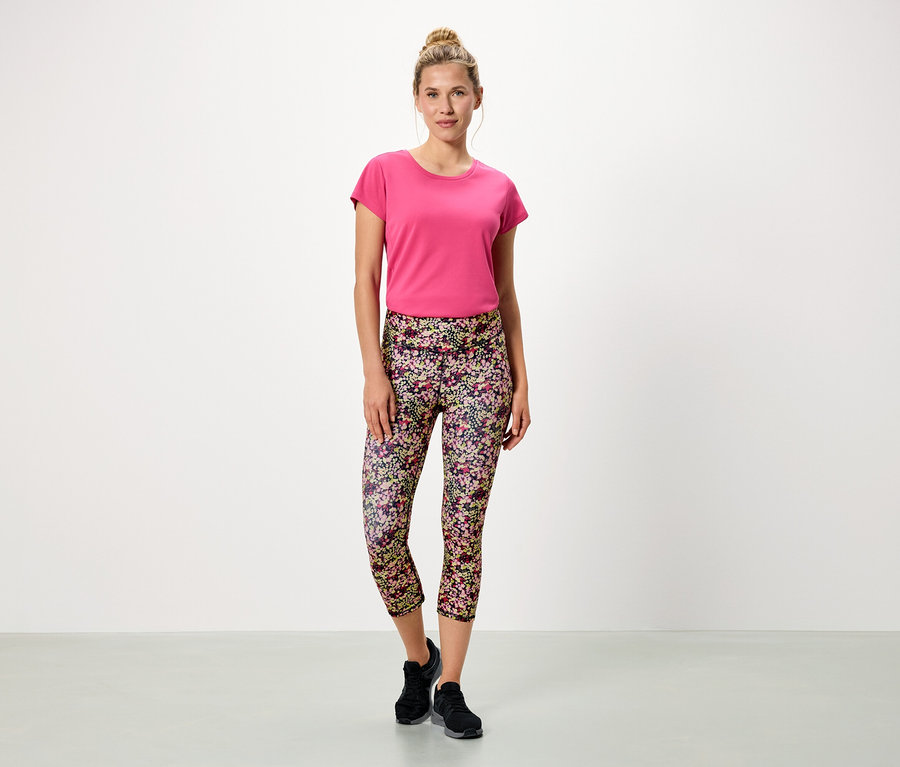 Žena stojí na bílém pozadí v růžovém tričku a květinových legínách.