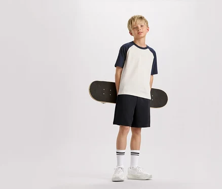 Blonďatý chlapec drží skateboard za zády, má na sobě tričko s tmavě modrými rukávy, šortky a tenisky.