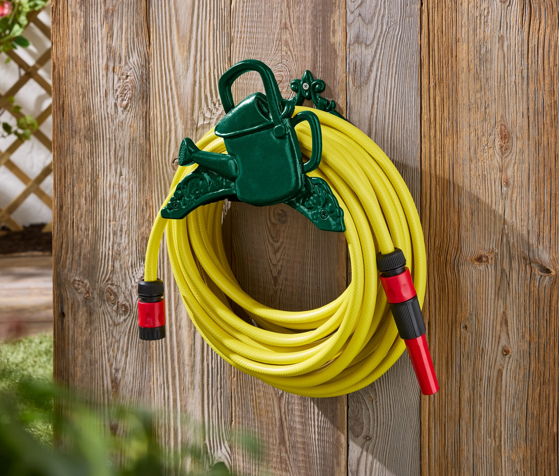 Žlutá zahradní hadice připevněná k zelenému držáku ve tvaru konve na dřevěné stěně.