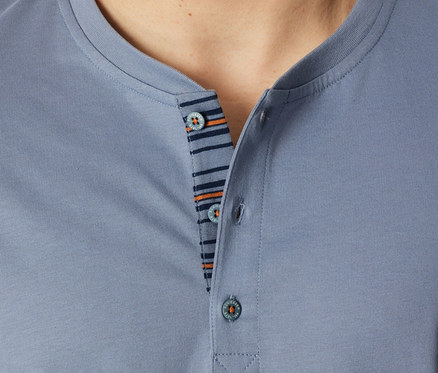 Detailní záběr osoby v modrošedém topu z krátké pyžamové soupravy SCHIESSER, ukazující Henley výstřih se třemi knoflíky a pruhovanou légu v tmavě modré a oranžové barvě.