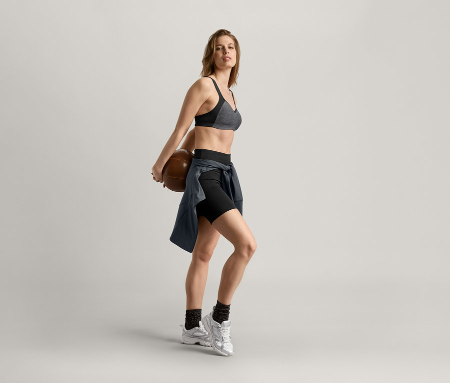 Žena drží za zády basketbalový míč, má na sobě sportovní podprsenku, šortky a sportovní bundu antracitové barvy.