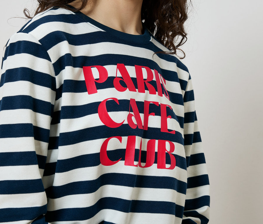 Žena má na sobě modrobílou pruhovanou mikinu s červeným nápisem „Paris Cafe Club“.