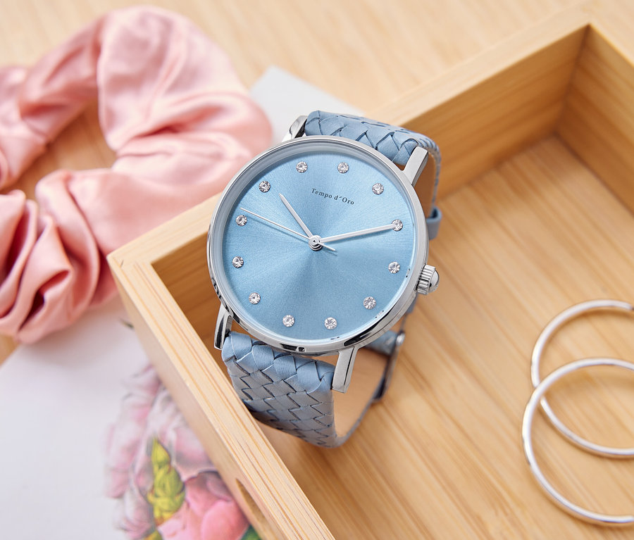 Světle modré hodinky se štrasovými kamínky leží v dřevěné krabičce vedle růžového scrunchie, zápisníku a stříbrných kruhů.