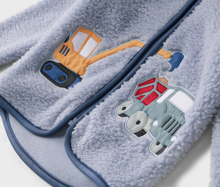 Detailní pohled na světle modrou fleecovou bundu se zipem a našitými motivy bagru a náklaďáku.