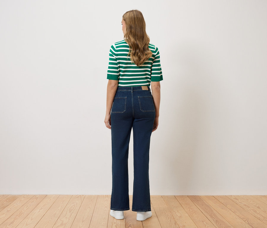 Pohled zezadu na ženu s dlouhými vlnitými vlasy v zeleno-bílém pruhovaném topu a tmavě modrých džínách s rozšířenými nohavicemi a bílými teniskami.