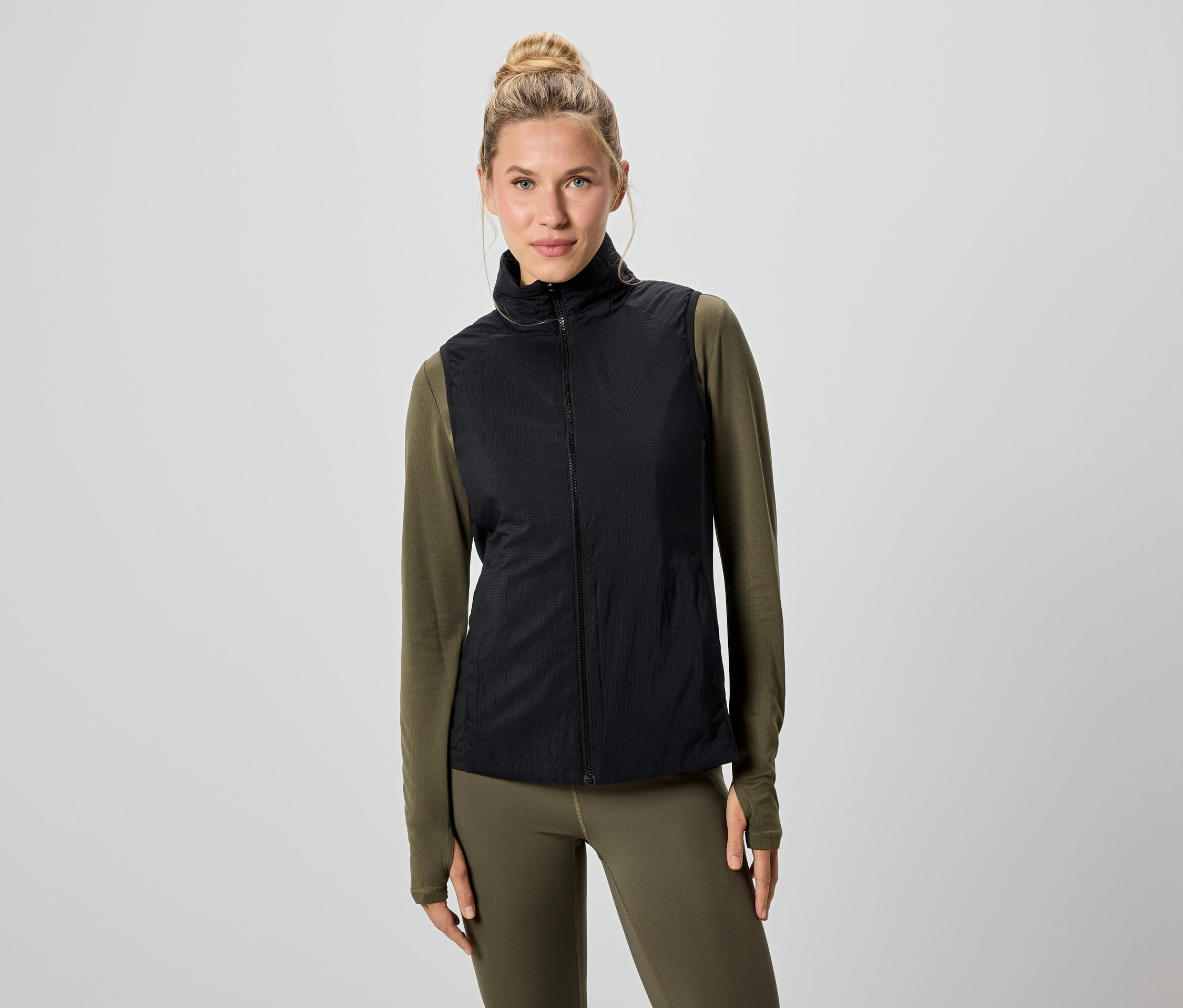 Žena s blond vlasy má na sobě černou vestu přes zelené tričko s dlouhým rukávem a legíny.